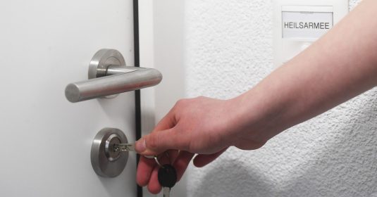 Eine Frauenhand steckt einen Schlüssel in die Wohnungstür, die mit Heilsarmee angeschrieben ist.