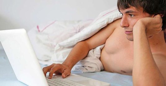 Mann, der mit nacktem Oberkörper im Bett liegt. Vor ihm ist ein Laptop.