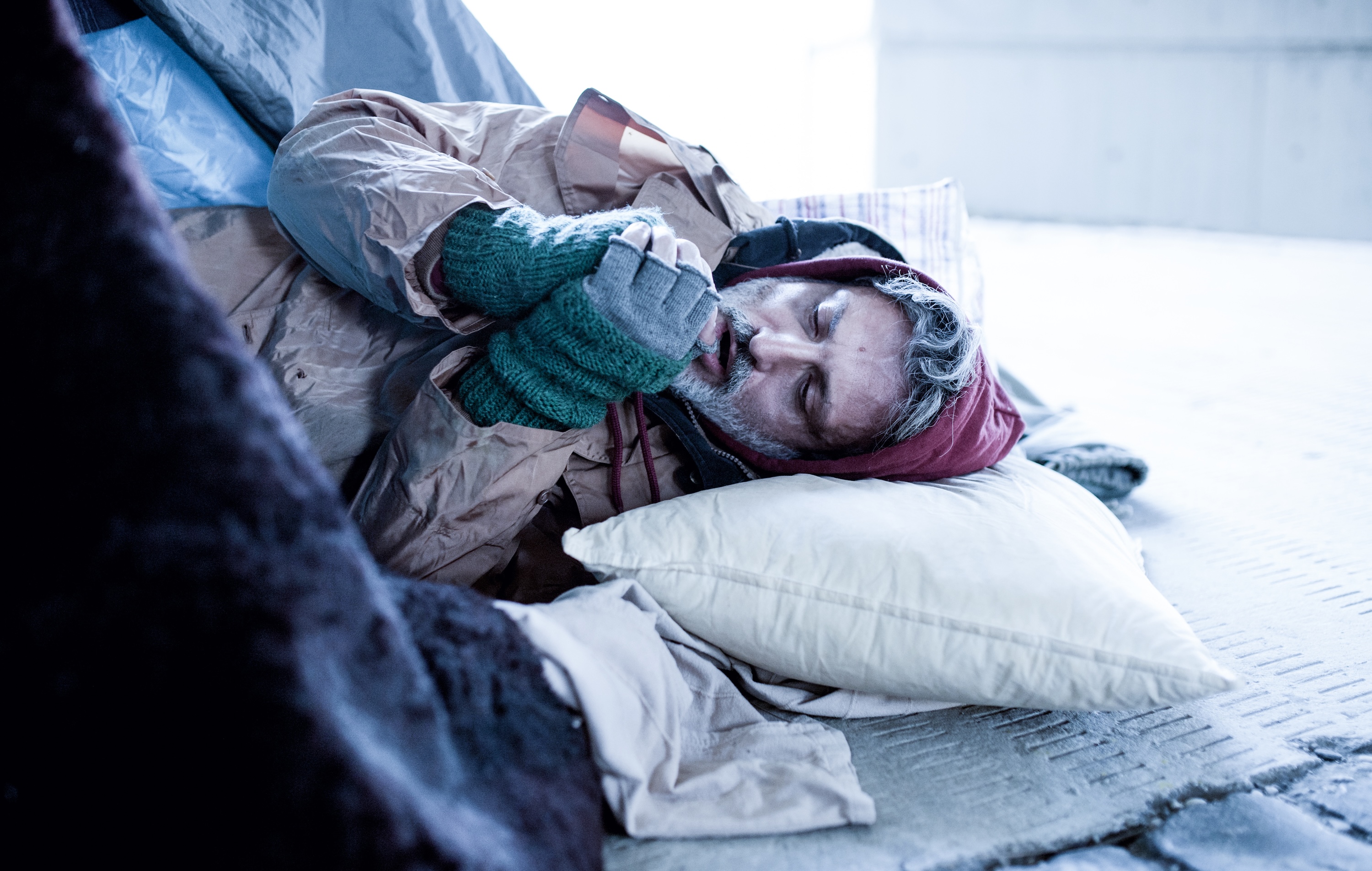 Homeless beggar man lying on the ground outdoors in city, warming up hands.